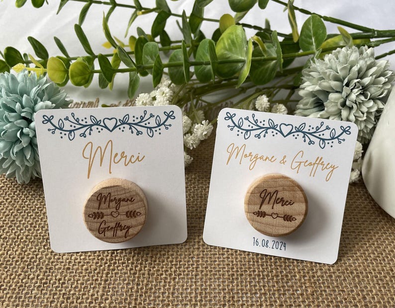 May include: Two white cards with a floral design and the word "Merci" in gold script. Each card has a small, round wooden magnet with engraved text. One magnet says "Morgane & Geoffrey" and the other says "Merci".