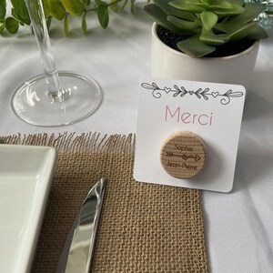 May include: A place card with the word "Merci" in pink, a wooden disc with names and an arrow design, and a small succulent plant in a white pot. The place card is on a burlap placemat.