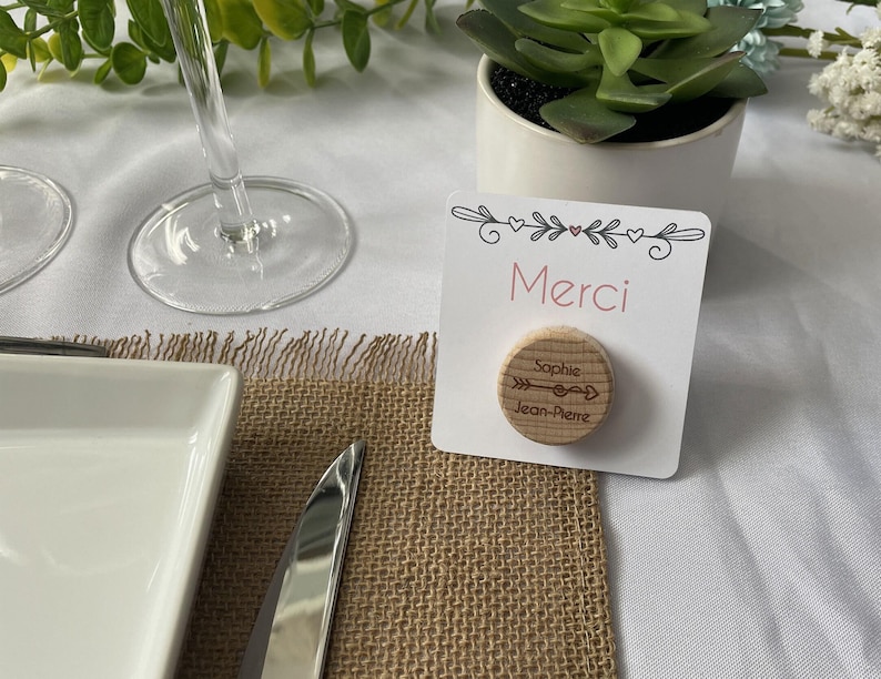 May include: A place card with the word "Merci" in pink, a wooden disc with names and an arrow design, and a small succulent plant in a white pot. The place card is on a burlap placemat.
