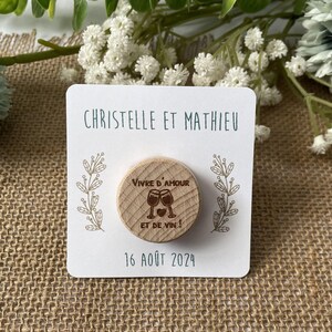 May include: A wooden magnet with the text "Vivre d'amour et de vin!" and two wine glasses. The magnet is on a white card with the names "Christelle et Mathieu" and the date "16 ao&ucirc;t 2024".