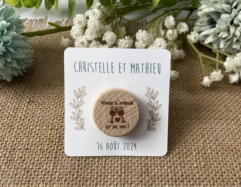 May include: A wooden magnet with the text "Vivre d'amour et de vin!" and two wine glasses. The magnet is on a white card with the names "Christelle et Mathieu" and the date "16 ao&ucirc;t 2024".