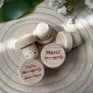 May include: A collection of wooden bottle stoppers with corks. Two stoppers have the names "Sophie Jean-Pierre" and the word "Merci" engraved with an arrow design. The stoppers are light brown and sit on a light brown wooden surface.