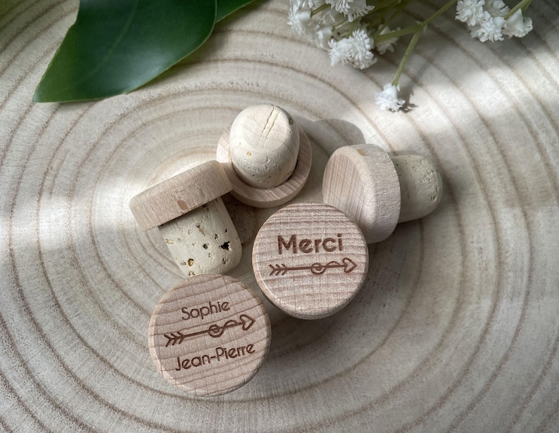 May include: A collection of wooden bottle stoppers with corks. Two stoppers have the names "Sophie Jean-Pierre" and the word "Merci" engraved with an arrow design. The stoppers are light brown and sit on a light brown wooden surface.