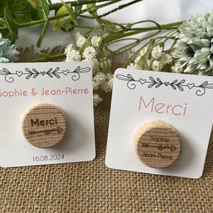 May include: Two white cards with pink text and wooden circles. One card says "Sophie & Jean-Pierre" and "16.08.2024". The other says "Merci". The wooden circles have the word "Merci" and an arrow design.