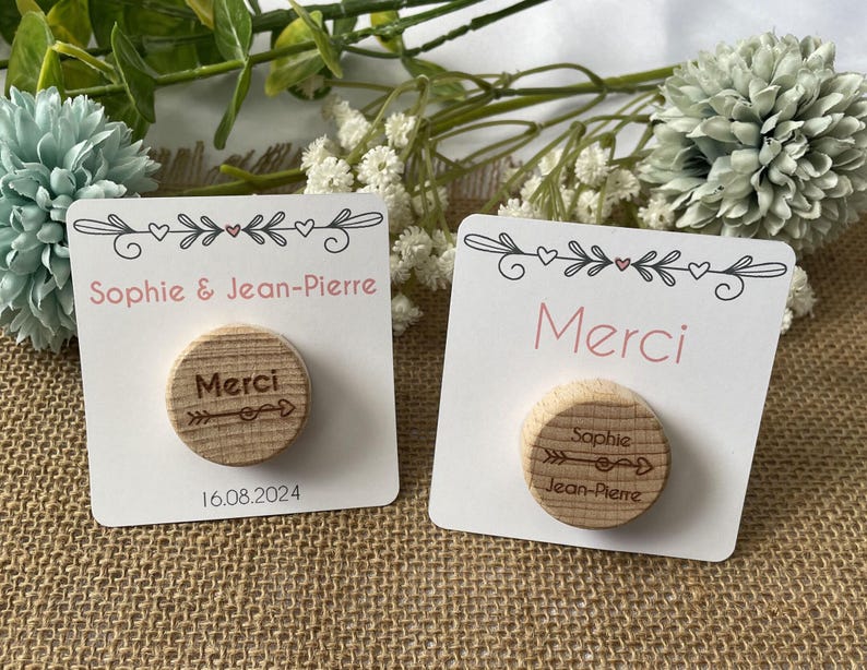 May include: Two white cards with pink text and wooden circles. One card says "Sophie & Jean-Pierre" and "16.08.2024". The other says "Merci". The wooden circles have the word "Merci" and an arrow design.