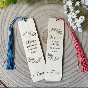 Peut inclure: Deux marque-pages en bois avec des pompons. L'un a un pompon bleu, l'autre un pompon rose. Les deux portent les mots "MERCI maitresse pour cette année" et "MERCI à mon ATSEM pour cette année" gravés, avec des motifs floraux et le nom "Leo" et des dates.