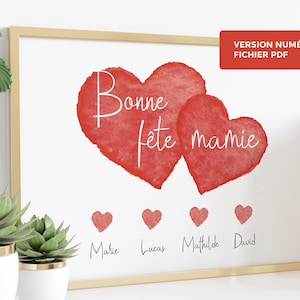 Op de afbeelding: Een ingelijste print met een gouden rand toont twee rode aquarel harten en de woorden "Bonne fête mamie" in witte letters. Daaronder staan vier kleine rode harten met namen. Rechtsboven staat de tekst: "VERSION NUMÉRIQUE FICHIER PDF".