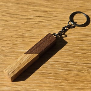 Könnte beinhalten: Ein Holzschlüsselanhänger mit rechteckigem Design. Die obere Hälfte ist dunkelbraun, während die untere Hälfte einen helleren, natürlichen Holzton aufweist. Der Schlüsselanhänger hat einen silberfarbenen Ring und eine Kette.