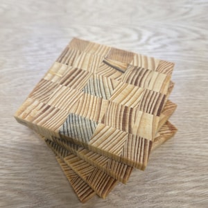 Peut inclure: Une pile de quatre dessous de verre carrés en bois. Chaque dessous de verre est fait de petits morceaux de bois rectangulaires disposés en motif géométrique. Le bois est de couleur brun clair naturel avec un grain visible.