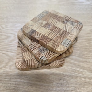 May include: Four square wooden coasters stacked on top of each other. Each coaster is made from small, rectangular pieces of light and dark brown wood, arranged in a checkerboard pattern. The edges are rounded, and they rest on a light-coloured wooden surface.