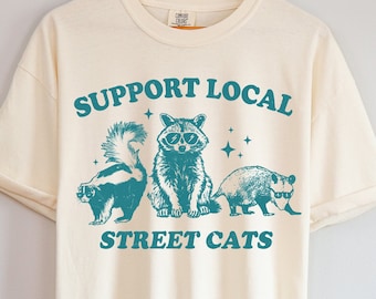 May include: A cream colored t-shirt with a teal graphic of a raccoon, skunk, and opossum wearing sunglasses. The text "Support Local Street Cats" is printed on the shirt.