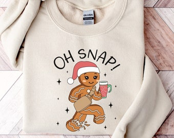 Könnte beinhalten: Ein beiger Pullover mit einem Lebkuchenmann-Motiv und dem Text "OH SNAP!" auf der Vorderseite. Der Lebkuchenmann trägt eine rote Weihnachtsmütze und hält ein Glas Eierlikör.