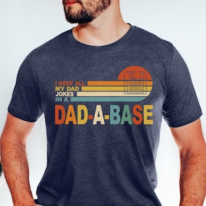 May include: Heather navy t-shirt with the text "I KEEP ALL MY DAD JOKES IN A DAD-A-BASE" in a retro design. The graphic includes a sunset-style background with orange, yellow, and green stripes. The shirt is a crew neck.