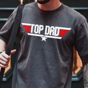May include: Dark gray t-shirt with the text "TOP DAD" in white, with red wing accents and a star. The shirt is a crew neck style and has a vintage, worn look.
