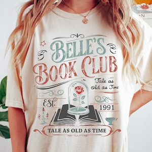 May include: Ivory-colored t-shirt with a graphic design for Belle's Book Club. The design includes text, a rose in a glass dome, and the phrase "Tale as old as Time." The shirt also has the year "1991" printed on it.