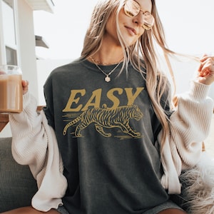 May include: Dark gray sweatshirt with the word "EASY" in gold lettering above a gold tiger illustration. The sweatshirt is worn with gray shorts and a white cardigan. The word "PEPPER" is at the bottom of the image.