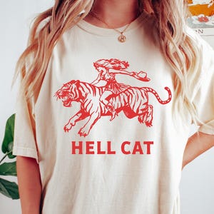 May include: A white t-shirt with a red graphic of a woman riding a tiger. The text "HELL CAT" is printed below the graphic. The shirt is available in ivory.