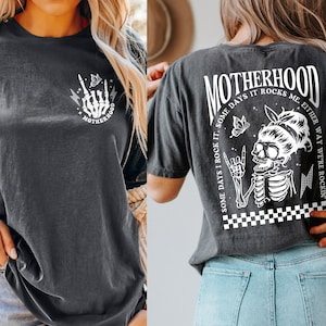 May include: A black t-shirt with a white graphic print on the front and back. The front graphic is a skeleton hand with a lightning bolt and the word 'MOTHERHOOD'. The back graphic is a skeleton with a butterfly and the text 'MOTHERHOOD SOME DAYS I ROCK IT. SOME DAYS IT ROCKS ME. EITHER WAY WE'RE ROCKIN''.