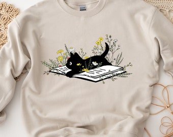 May include: A beige crewneck sweatshirt featuring a graphic print of a black cat lying on an open book surrounded by yellow and white wildflowers. The design is centered on the chest of the garment, which has long sleeves and a ribbed neckline.