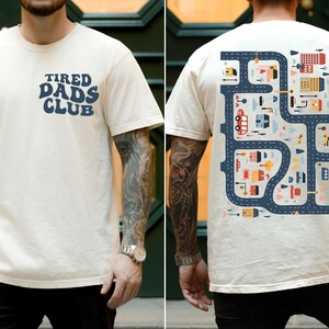 K&ouml;nnte beinhalten: Elfenbeinfarbenes T-Shirt mit dem Text "TIRED DADS CLUB" in Marineblau auf der Vorderseite. Die R&uuml;ckseite zeigt ein Stra&szlig;enkarten-Design mit farbenfrohen Geb&auml;uden, Fahrzeugen und Stra&szlig;en. Das T-Shirt ist kurz&auml;rmelig.