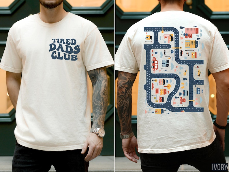 K&ouml;nnte beinhalten: Elfenbeinfarbenes T-Shirt mit dem Text "TIRED DADS CLUB" in Marineblau auf der Vorderseite. Die R&uuml;ckseite zeigt ein Stra&szlig;enkarten-Design mit farbenfrohen Geb&auml;uden, Fahrzeugen und Stra&szlig;en. Das T-Shirt ist kurz&auml;rmelig.