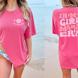 May include: A bright pink short-sleeve t-shirt featuring a small light pink smiley face graphic with lightning bolts and the text "GIRL MAMA ERA" on the front left chest, and a large matching graphic on the back with the text "IN MY GIRL MAMA ERA".
