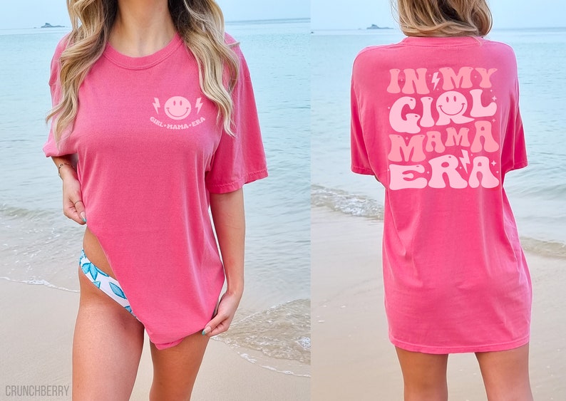 May include: A bright pink short-sleeve t-shirt featuring a small light pink smiley face graphic with lightning bolts and the text "GIRL MAMA ERA" on the front left chest, and a large matching graphic on the back with the text "IN MY GIRL MAMA ERA".
