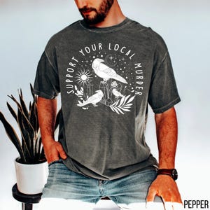 May include: Dark grey t-shirt with a white graphic that reads "Support Your Local Murder." The graphic features three stylized birds, a sun, moon, stars, and geometric shapes. The shirt is a relaxed fit.