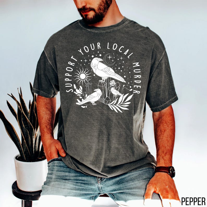 May include: Dark grey t-shirt with a white graphic that reads "Support Your Local Murder." The graphic features three stylized birds, a sun, moon, stars, and geometric shapes. The shirt is a relaxed fit.