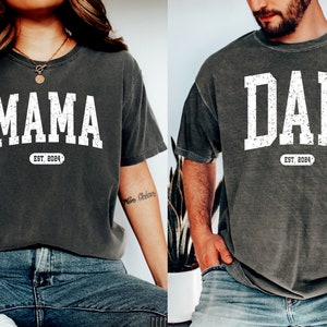 May include: Two gray t-shirts with white text. The shirt on the left says "MAMA EST. 2024" and the shirt on the right says "DAD EST. 2024".