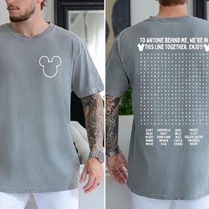 May include: Grey t-shirt with a white Mickey Mouse outline on the front. The back of the shirt features a word search puzzle with Disney-themed words and the text "TO ANYONE BEHIND ME, WE'RE IN THIS LINE TOGETHER. ENJOY!"