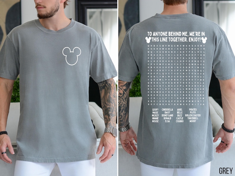 May include: Grey t-shirt with a white Mickey Mouse outline on the front. The back of the shirt features a word search puzzle with Disney-themed words and the text "TO ANYONE BEHIND ME, WE'RE IN THIS LINE TOGETHER. ENJOY!"