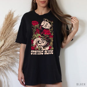 May include: Black t-shirt with a graphic of a skull with coffee pouring into its mouth, surrounded by red roses and vines. The text "STAYING ALIVE" is printed below the skull.