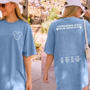 May include: A blue jean colored t-shirt with a white Mickey Mouse outline on the front. The back of the shirt features the text "TO ANYONE BEHIND ME, WE'RE IN THIS LINE TOGETHER. ENJOY!" and a word search with Disney-related words.