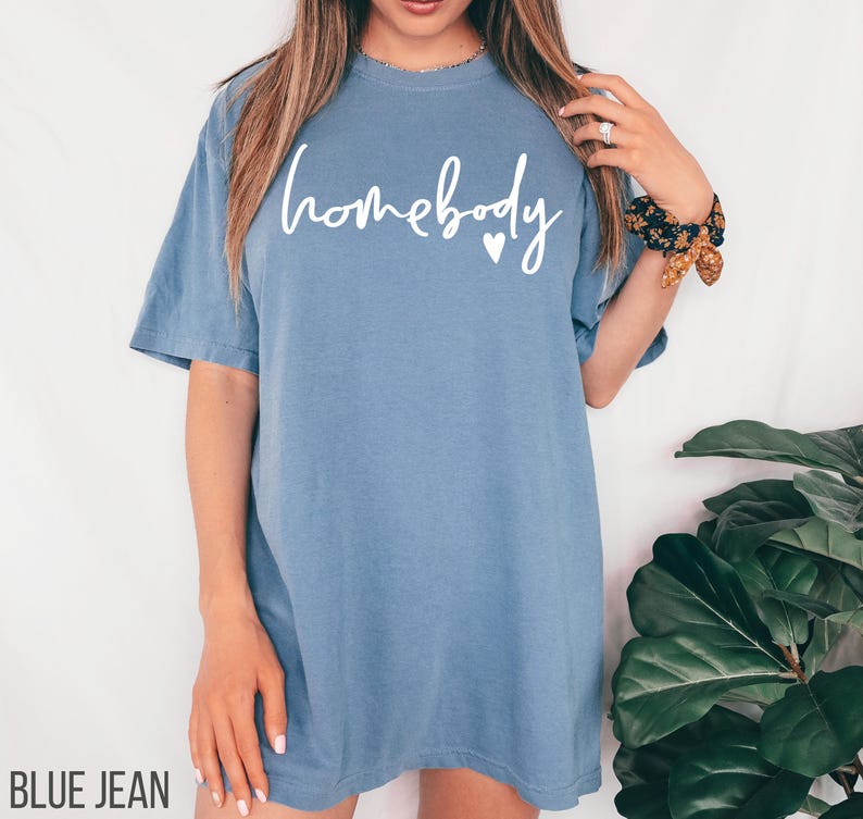 Puede incluir: Una camiseta de gran tama&ntilde;o de color azul jean con el texto "homebody" en letras blancas y un peque&ntilde;o coraz&oacute;n.