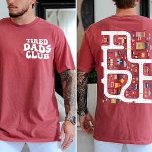 K&ouml;nnte beinhalten: Karmesinrotes T-Shirt mit dem wei&szlig;en Schriftzug "TIRED DADS CLUB" auf der Vorderseite. Die R&uuml;ckseite zeigt ein farbenfrohes Stra&szlig;enkarten-Design mit Geb&auml;uden und Fahrzeugen. Ein l&auml;ssiges, bequemes T-Shirt.