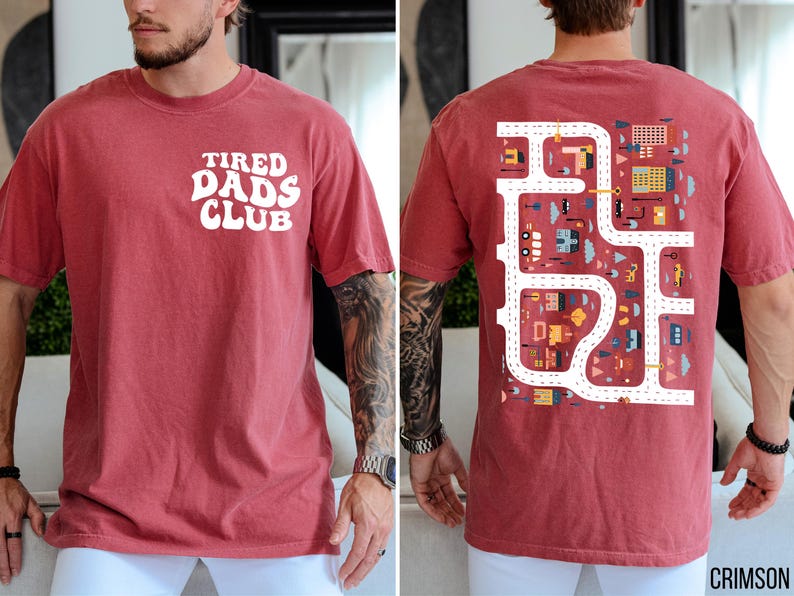 K&ouml;nnte beinhalten: Karmesinrotes T-Shirt mit dem wei&szlig;en Schriftzug "TIRED DADS CLUB" auf der Vorderseite. Die R&uuml;ckseite zeigt ein farbenfrohes Stra&szlig;enkarten-Design mit Geb&auml;uden und Fahrzeugen. Ein l&auml;ssiges, bequemes T-Shirt.