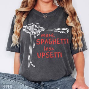 May include: A dark gray t-shirt with a white graphic of a fork with spaghetti on it. The text "more spaghetti less upsetti" is written in white with the word "upsetti" in red.