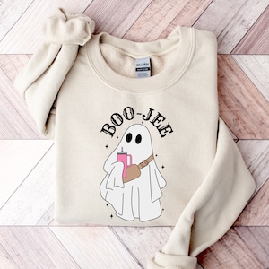 May include: A beige sweatshirt featuring a white ghost holding a pink drink and a brown strap across its chest. The text "BOO-JEE" is written in black above the ghost.