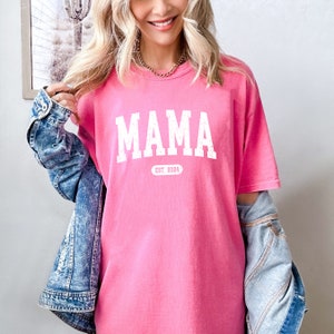 May include: Pink t-shirt with white text that reads "MAMA EST. 2024".