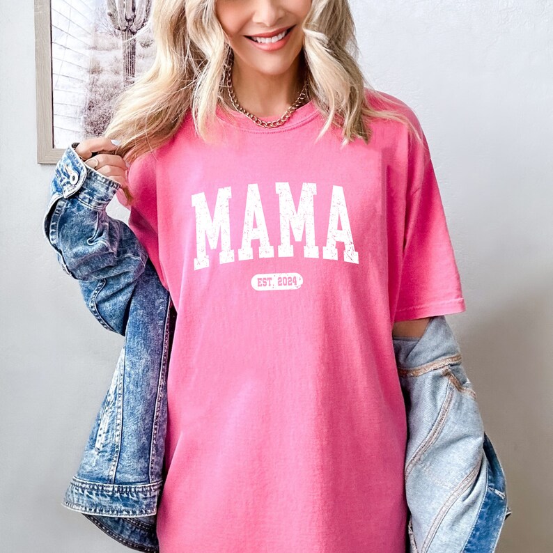 May include: Pink t-shirt with white text that reads "MAMA EST. 2024".