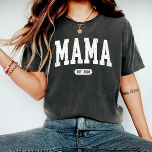 May include: A dark gray t-shirt with the word "MAMA" printed in white with a distressed look. Below the word is a white oval with the text "EST. 2024" inside.