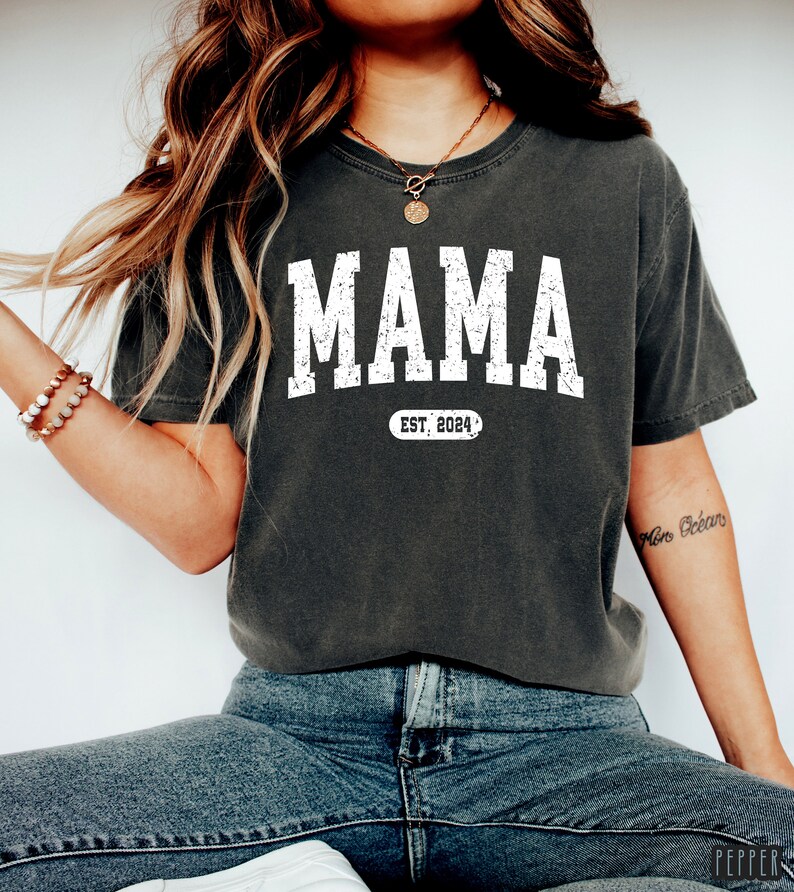 May include: A dark gray t-shirt with the word "MAMA" printed in white with a distressed look. Below the word is a white oval with the text "EST. 2024" inside.