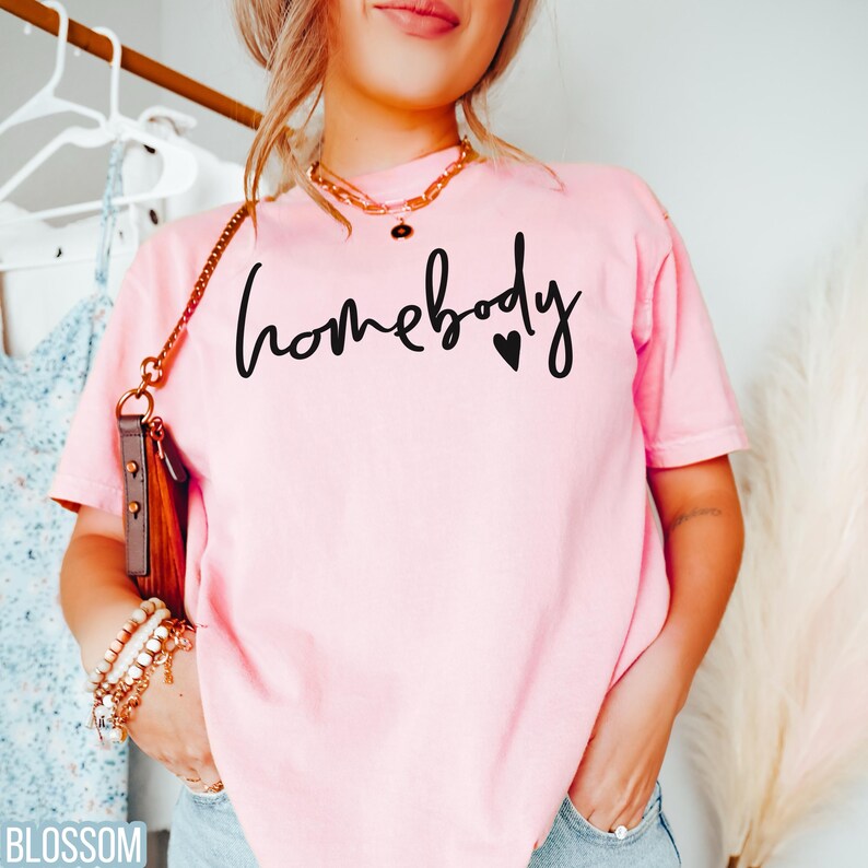Puede incluir: Una camiseta rosa con el texto "homebody" en letra negra con un peque&ntilde;o coraz&oacute;n negro.