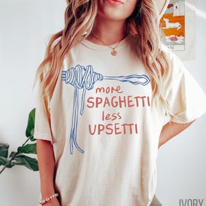 May include: A cream colored t-shirt with a blue and red graphic of a fork with spaghetti on it. The text on the shirt reads "more spaghetti less upsetti".