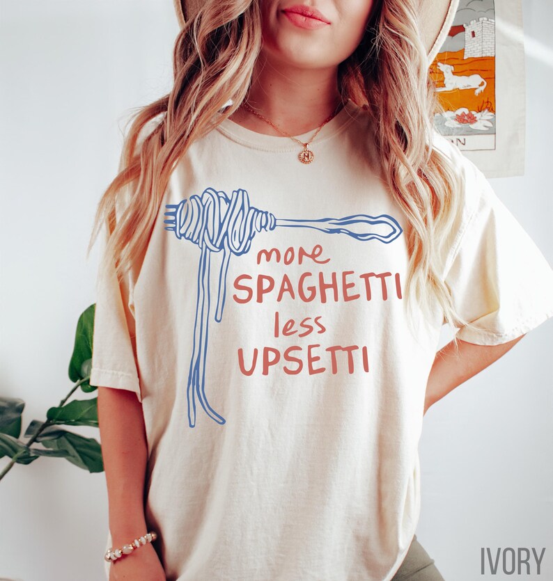 May include: A cream colored t-shirt with a blue and red graphic of a fork with spaghetti on it. The text on the shirt reads "more spaghetti less upsetti".