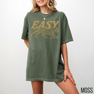 May include: Moss green t-shirt with the word "EASY" in gold lettering above a gold tiger graphic. The shirt is a relaxed fit and has short sleeves. The word "MOSS" is printed in the bottom right corner.