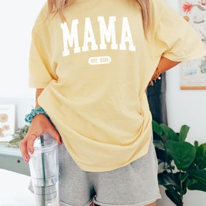 May include: A yellow t-shirt with the word "MAMA" printed in white with a distressed look. Below the word "MAMA" is a white oval with the text "EST. 2024" inside.