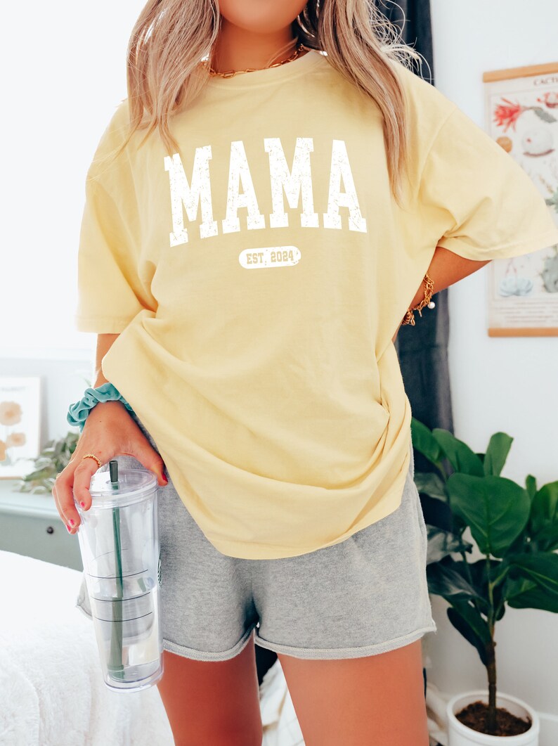 May include: A yellow t-shirt with the word "MAMA" printed in white with a distressed look. Below the word "MAMA" is a white oval with the text "EST. 2024" inside.