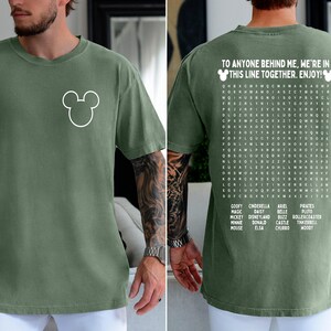 May include: Moss green t-shirt with a white Mickey Mouse outline on the front. The back of the shirt features a word search puzzle with Disney-themed words and the text "TO ANYONE BEHIND ME, WE'RE IN THIS LINE TOGETHER. ENJOY!"